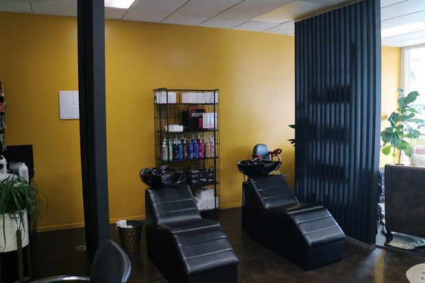 Interior view of a professional hair salon with two black reclining shampoo stations and integrated basins, a product shelving unit, dark vertical slatted partition, mustard-yellow walls, and polished dark flooring. Daylight enters through a window at right, with salon furnishings visible beyond the partition.