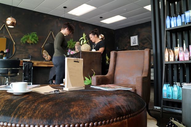 A stylish hair salon reception and waiting area with a dark, textured interior, leather seating, product shelving, pendant lighting, and staff assisting clients at the counter. A large tufted leather ottoman or table dominates the foreground, with salon products displayed on open shelving.