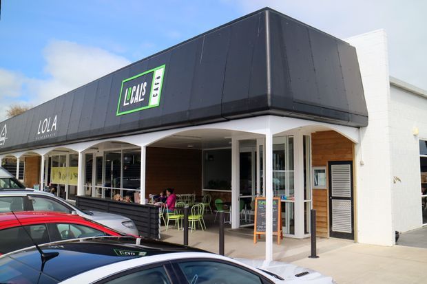 Street-level exterior view of a contemporary low-rise commercial retail and hospitality building. The dark charcoal fascia carries Lola and Lola Cafe signage, with a covered verandah, glazed storefronts, outdoor dining tables and chairs, timber-lined entrance areas, and white masonry side walls. Parked cars occupy the foreground.