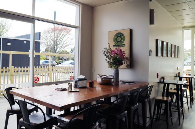 A bright casual dining area with a long solid timber communal table, black chairs and stools, and a narrow counter along the wall. Large windows provide views to the street, while framed artwork and floral arrangements add visual interest. The space has a clean, contemporary café or hospitality fit-out with pale walls and a suspended ceiling.