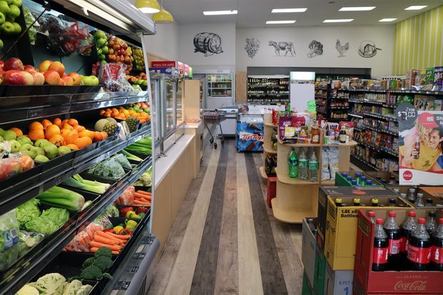 A brightly lit convenience grocery store interior with fresh produce displayed in refrigerated shelving along the left wall, central aisles stocked with packaged foods and beverages, checkout fixtures, promotional displays, and wood-look flooring. The space has a practical retail layout, white ceiling tiles with recessed lighting, and a striped green feature wall.
