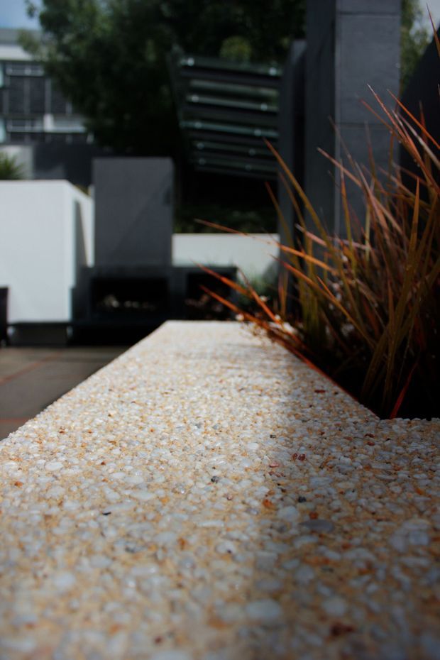 Close-up exterior view of a light pebble-textured concrete or aggregate planter wall/bench beside ornamental grasses. A dark contemporary facade, gate, and neighbouring residential forms are visible in the blurred background.