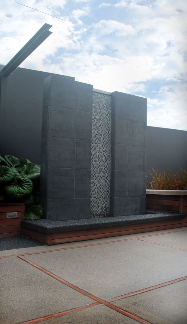 A modern outdoor courtyard or terrace feature with tall dark grey concrete-clad panels, a central strip of small mosaic tiles, and a shallow black pebble-textured water feature or planter base. Timber-edged raised planters with ornamental foliage flank the structure, while large pale concrete paving slabs with narrow terracotta-coloured joints cover the foreground. The composition is minimalist and geometric beneath a bright sky.