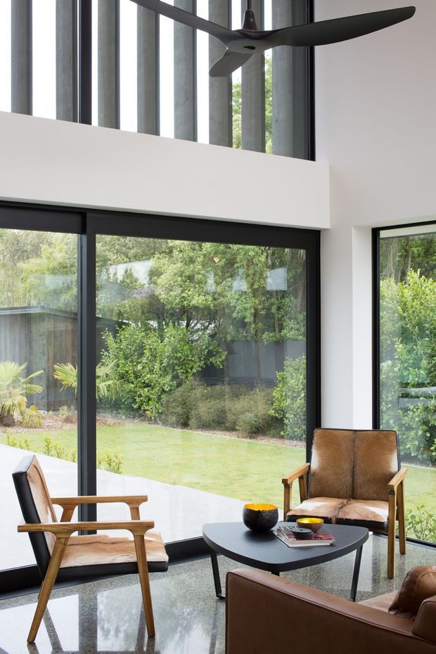 A bright, double-height living room with expansive black-framed glazing overlooking a landscaped garden. The room features polished grey flooring, white walls, a sculptural black ceiling fan, and mid-century-inspired leather and timber lounge chairs arranged around a low dark coffee table.
