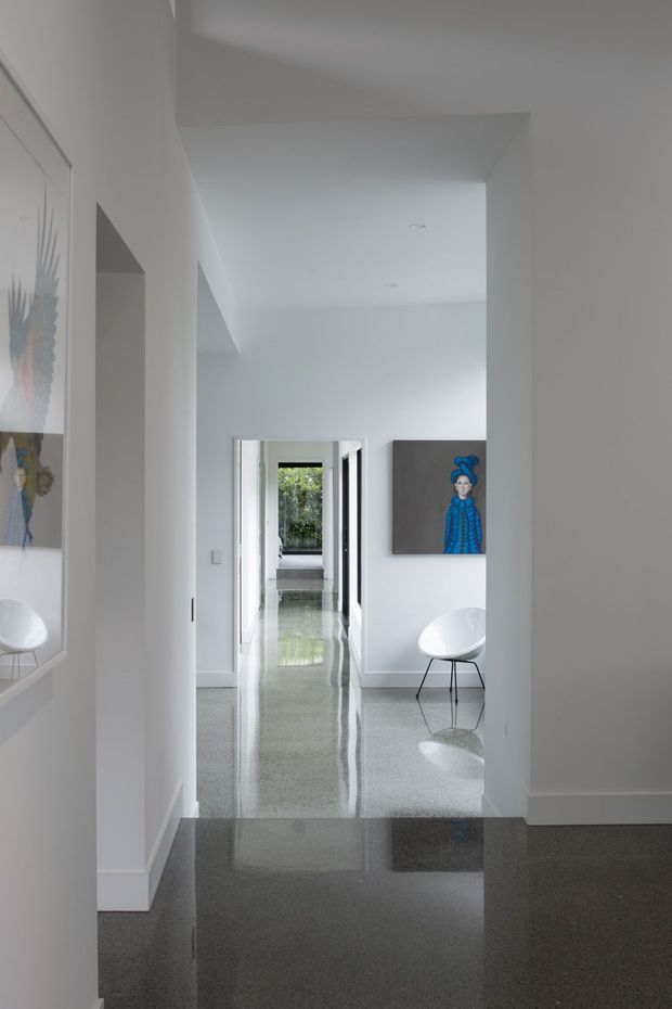 A minimalist, bright interior hallway viewed through a series of aligned openings. The space has crisp white walls and ceilings, a highly polished grey terrazzo floor with strong reflections, recessed downlights, and white door frames. A sculptural white chair and colourful artwork appear along the corridor, with greenery visible through a distant opening.