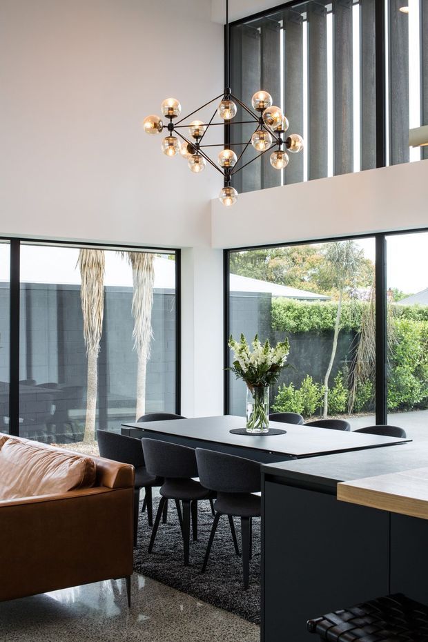 A sophisticated open-plan dining and kitchen interior with a double-height ceiling, expansive black-framed glazing, and a sculptural multi-bulb pendant light. A dark rectangular dining table is surrounded by upholstered black chairs, with a brown leather sofa at left and dark kitchen cabinetry with a light timber benchtop at right. Polished speckled flooring, white walls, and a landscaped courtyard visible through the windows create a refined contemporary atmosphere.