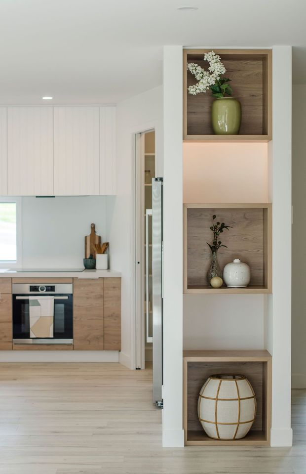 Bright contemporary kitchen and hallway interior with white flat-panel cabinetry, light timber flooring, pale timber lower cabinets, and integrated shelving niches. The built-in display shelving has warm timber backs, soft accent lighting, and decorative vases. A built-in oven and concealed rangehood are visible in the kitchen.