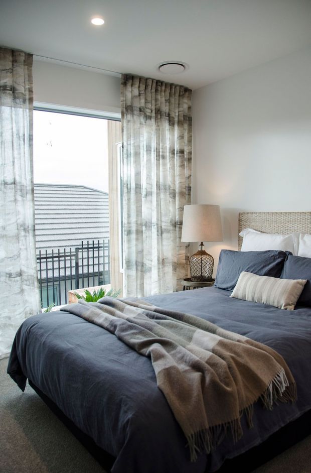 A calm, softly lit bedroom with a large bed dressed in deep blue-grey linens and layered neutral throws and pillows. The room features a woven natural-fibre headboard, sheer patterned curtains, a bedside table with a warm fabric lamp, recessed ceiling lighting, and a large glazed door or window opening onto a balcony with a neighbouring roof visible outside.
