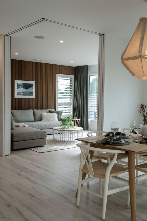 A bright, refined open-plan interior showing a dining area in the foreground and a living room beyond. The space features pale timber flooring, a natural timber dining table with white cross-back chairs, a grey sectional sofa, a round wire-frame coffee table, and a dark vertical timber feature wall. Large sliding partition doors, plantation shutters, curtains, framed artwork, and a sculptural woven pendant light complete the calm contemporary setting.