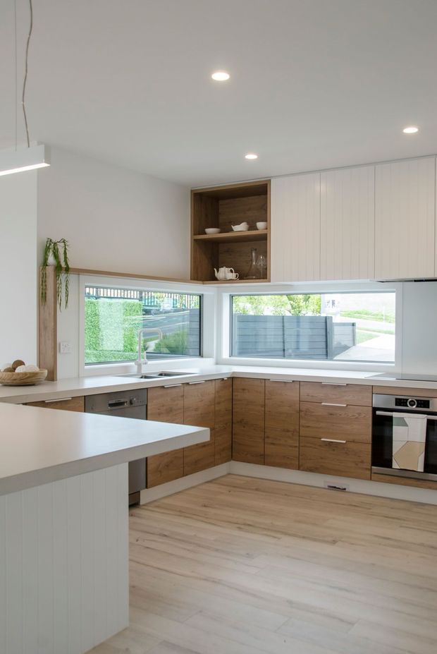 Bright contemporary kitchen with an L-shaped run of warm wood cabinetry, white benchtops, integrated appliances, and a white island with vertically panelled sides. Large corner windows provide natural light, while open timber shelving displays crockery. Pale timber-look flooring and recessed ceiling lighting create a clean, inviting interior.