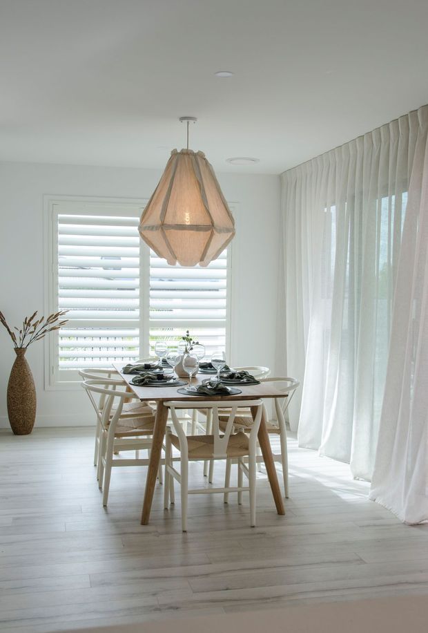 Bright, airy dining room with a light timber dining table and matching woven-back chairs, set with dark plates and glassware. White plantation shutters, sheer full-height curtains, pale timber flooring, and a large woven pendant light create a soft coastal-inspired atmosphere.