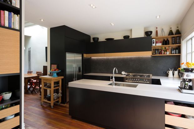 A contemporary open-plan kitchen with a large white island and integrated sink, matte black cabinetry, dark stone or composite splashback, stainless-steel appliances, and open timber shelving. Warm timber flooring and adjacent dining furniture add contrast to the monochrome cabinetry. Recessed ceiling lighting provides a bright, clean atmosphere.