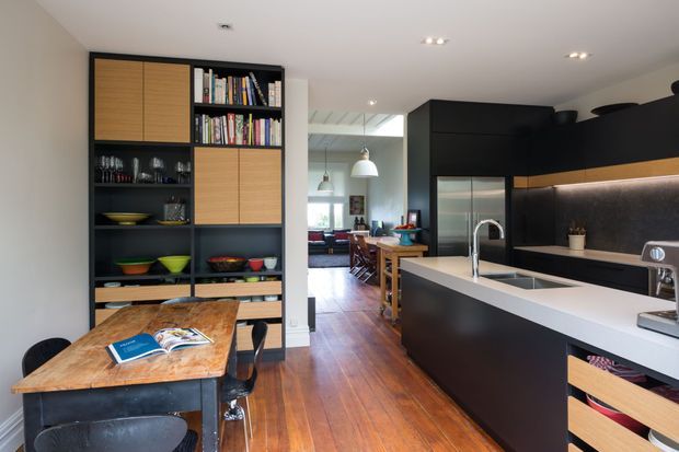 Open-plan contemporary kitchen and dining area with dark matte cabinetry, a pale stone island benchtop, integrated appliances, timber flooring, and a built-in shelving unit with timber-fronted cupboards. A dining table sits beside the kitchen, with a living area visible beyond.