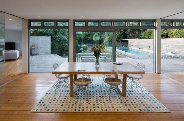 A bright, open-plan dining area with a long timber dining table and woven wire-frame chairs arranged on a patterned rug. Full-height glazed doors and clerestory windows open the room visually to a timber deck, landscaped garden, and rectangular swimming pool. Warm timber flooring, dark-framed glazing, and a minimalist built-in interior create a clean indoor–outdoor connection.