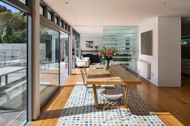 Bright contemporary dining area with a long timber dining table and wire-frame chairs positioned beside full-height glazing. Warm timber flooring, a patterned area rug, white walls, recessed ceiling lights, and a translucent horizontal glass partition define the open-plan space. A living area is visible beyond, while the glazing opens toward an exterior deck and pool area.