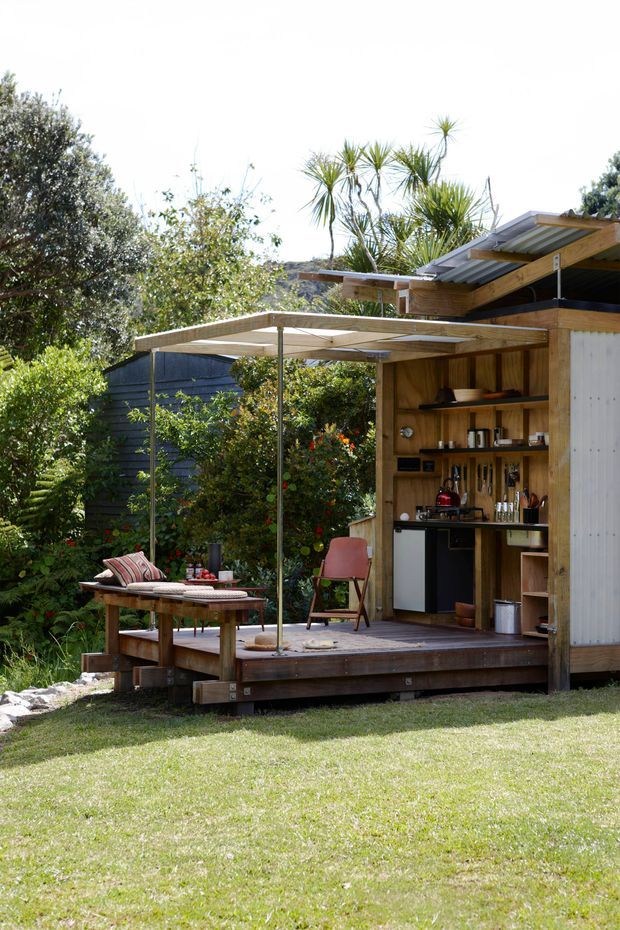 A compact timber outdoor pavilion set within a lush garden, with a raised timber deck, covered dining/lounge area, and an integrated open-shelved kitchenette or bar. The structure has warm natural wood cladding, corrugated metal roofing, a simple flat canopy, and a relaxed garden setting with mature vegetation.