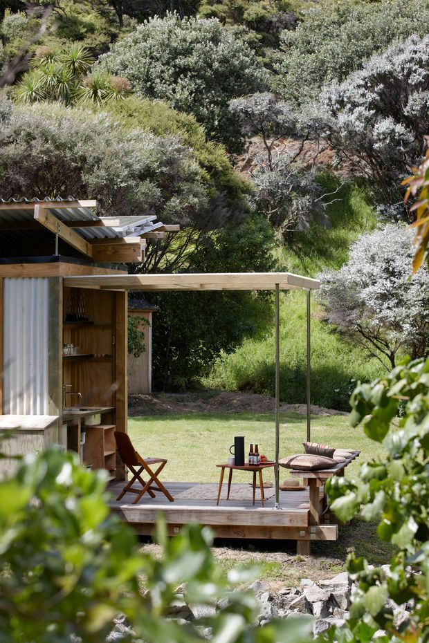 A compact timber outdoor pavilion or cabin set within dense native vegetation. The structure has corrugated metal roofing, timber walls and decking, a simple covered terrace with slender posts, and an outdoor seating area with a small table and chairs. The landscaped setting is leafy, informal, and strongly connected to the surrounding bush.