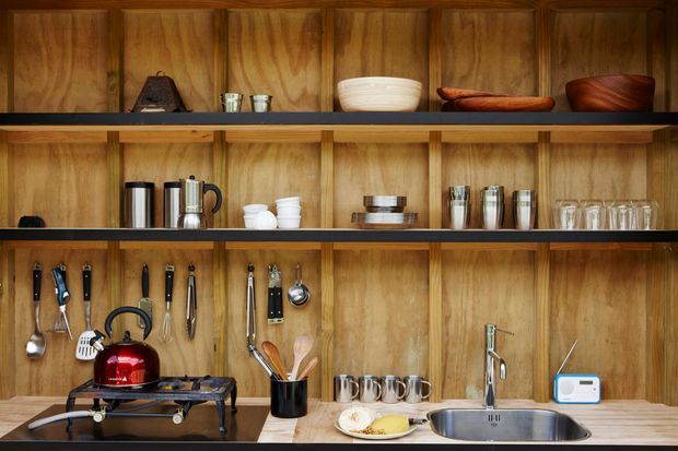 Close-up view of a warm timber-lined kitchen wall with open shelving, displaying cookware, crockery, utensils, small appliances, and glassware. A timber benchtop runs along the bottom with a freestanding gas burner, red kettle, utensil holder, and stainless-steel sink with tap. The composition has strong linear shelving and wood panelling, creating a practical yet curated kitchen workspace.