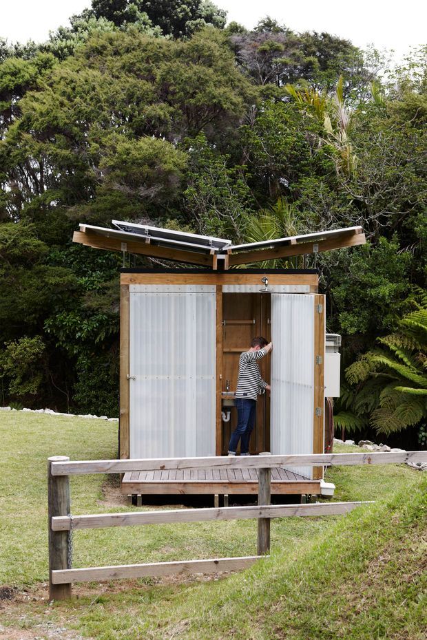 A compact freestanding outdoor shower structure set in a lush garden. The small timber-framed enclosure has translucent corrugated wall panels, an open central shower area with visible plumbing, and a simple split-pitched roof with exposed timber construction. It stands on a raised timber deck behind a rustic timber fence, surrounded by dense greenery and lawn.