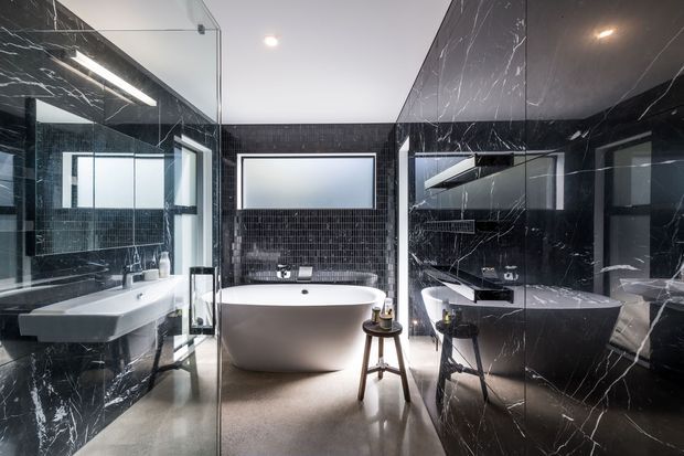 Luxurious contemporary bathroom with dramatic black marble-look wall surfaces, glossy dark mosaic tilework, a freestanding white bathtub, wall-mounted basin, frameless glass shower enclosure, large mirror and polished neutral flooring. The symmetrical composition and reflective finishes create a high-contrast, sophisticated atmosphere.