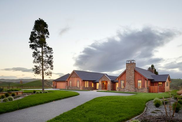 A sprawling single-storey rural residence with multiple gabled wings, warm timber cladding, substantial natural-stone wall sections, and a prominent stone chimney. The home sits within manicured lawns and curved gravel landscaping, with rolling green hills and a tall mature tree in the surrounding setting. Exterior architectural photography in soft overcast evening light.