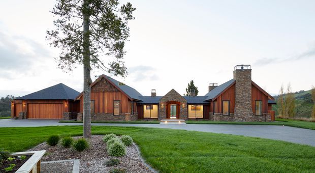 Wide exterior view of a sprawling single-storey rural residence with multiple pitched and gabled roof forms. The facade combines warm vertical timber cladding, natural stone masonry, dark roofing, and timber-framed windows and doors. A connected garage wing sits to the left, while a landscaped lawn, driveway, mature trees, and surrounding hills frame the property.