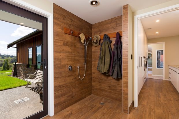 A contemporary mudroom-style entry with a walk-in shower or wash-down area finished in warm wood-look wall and floor surfaces. Hooks hold coats and hats, while a glazed exterior door opens to a lawn and garden. A bright hallway with built-in cabinetry is visible through the adjacent doorway.
