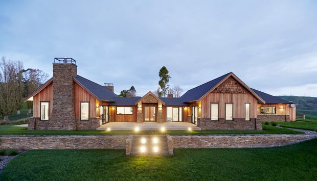 A wide exterior view of a low-slung, symmetrical residential home set in a landscaped rural setting. The house features multiple steep gabled wings, dark pitched roofing, warm timber-clad facades, extensive glazing, prominent rough-stone chimneys and masonry retaining walls. Illuminated steps lead to the central entrance, with open lawn and rolling hills beyond.