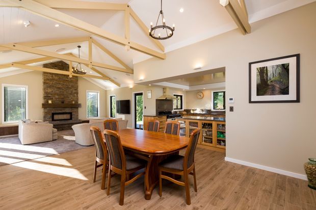 Open-plan dining and living area with a warm timber dining table and chairs in the foreground, a lounge with fireplace to the left, and a kitchen with island and timber cabinetry at the rear. The room has a vaulted ceiling with exposed pale timber trusses, light walls, timber-look flooring, abundant windows, and a central ring pendant light.