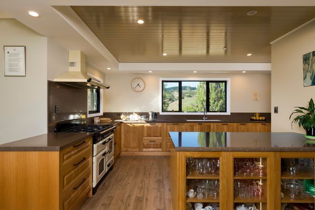 Warm, traditional-style kitchen with extensive timber cabinetry, a large freestanding range cooker, dark stone splashback, central island with a reflective dark benchtop, and a glazed display cabinet filled with glassware. A timber-lined recessed ceiling and views through the rear window add character and depth.