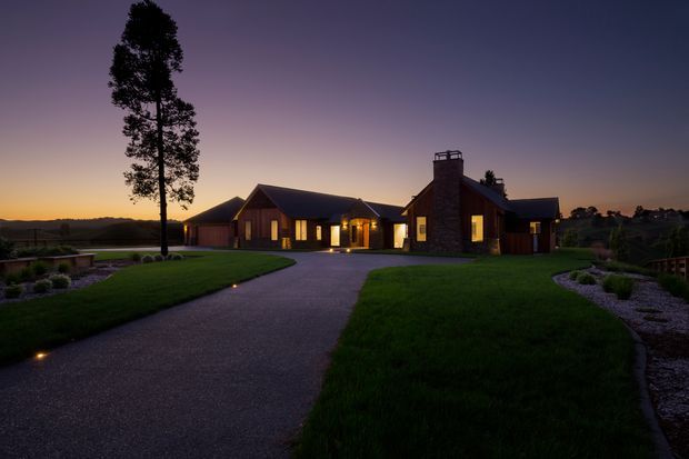 A sprawling single-storey residence photographed at dusk, approached by a broad curved driveway through expansive landscaped lawns. The home features dark pitched roofs, warm illuminated windows, a prominent stone-clad chimney, timber-toned wall sections, and multiple connected wings. Low landscape lighting and extensive planting frame the property against a rural-looking horizon.