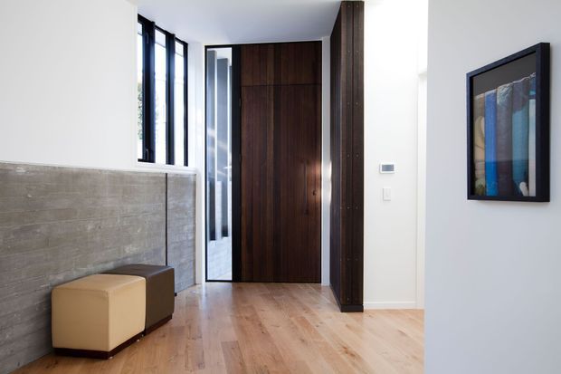 Bright contemporary entry hall with pale timber flooring, a tall dark timber front door and matching vertical joinery, a textured concrete half-height wall, narrow black-framed glazing, and simple cube ottomans. A framed abstract artwork adds a restrained blue accent on the white wall.