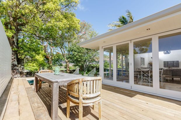 Bright outdoor timber deck adjoining a modern single-storey glazed living space. The deck features a weathered wooden dining table and chairs, white-framed full-height glass doors, a broad shallow roof eave, and leafy surrounding landscaping. The scene has a relaxed coastal or resort-like atmosphere, although the specific setting is not certain.