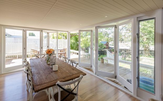 Bright enclosed dining sunroom with a large rustic timber dining table, mixed light chairs, pale timber flooring, a white slatted ceiling, and extensive floor-to-ceiling glazing with multiple glazed doors opening onto a deck and garden.