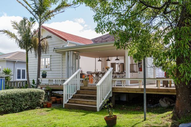 Exterior view of a raised timber-clad home with a covered outdoor deck and stair access. The house features horizontal weatherboard siding, a pitched red metal roof, white trim and balustrades, and an open entertaining area furnished with an outdoor dining table and pendant lights. Tropical planting and a manicured lawn surround the elevated structure.