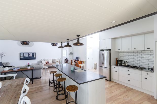 Bright open-plan kitchen and living area with white shaker-style cabinetry, a large dark stone island with timber stools, stainless-steel appliances, and a white subway-tile splashback. The adjoining living and dining areas are visible, with pendant lighting, pale timber flooring, and a white-lined ceiling.