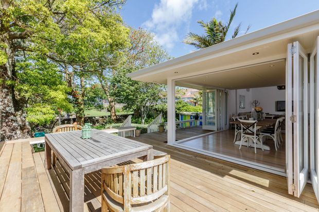 Sunny exterior view of a timber deck adjoining a contemporary open-plan living and dining area through fully opened glazed bi-fold doors. The deck has a large weathered timber dining table and chairs, while the interior features timber flooring, neutral walls, recessed ceiling lights, and casual dining furniture. Lush mature garden planting surrounds the deck, with a bright, airy indoor-outdoor connection.