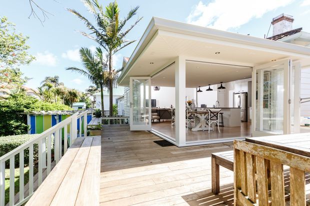 A bright, open-plan home extension opening onto a timber deck. The white-clad structure has extensive floor-to-ceiling glass doors, a covered outdoor area, and a visible kitchen and dining space inside. The deck includes built-in bench seating and a timber outdoor table, with tropical planting and a colourful fence visible beyond.