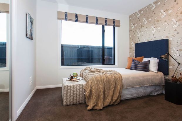A compact, softly styled bedroom with a single bed, navy upholstered headboard, layered neutral bedding, patterned wallpaper on the feature wall, dark carpet, and a large window with a striped Roman blind. A woven-texture ottoman sits at the foot of the bed, while a black bedside table and angled lamp add contrast.