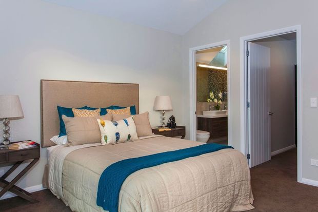 A softly styled bedroom with a large upholstered bed, layered beige bedding, and a vivid teal runner and cushions. Matching bedside tables and lamps flank the bed, while an open doorway reveals an adjacent bathroom with a vanity and patterned feature wall. The room has pale grey walls, carpeted flooring, white trim, and a calm contemporary aesthetic.