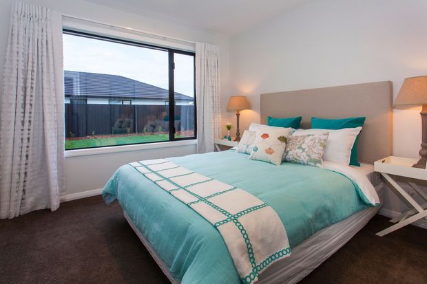 A bright, comfortably furnished bedroom with a large bed dressed in turquoise bedding, patterned accent cushions, and white curtains framing a wide window. Bedside tables with traditional lamps sit on either side, while dark carpet and neutral walls create a soft, coordinated palette. A neighboring roofline and fenced outdoor area are visible through the window.