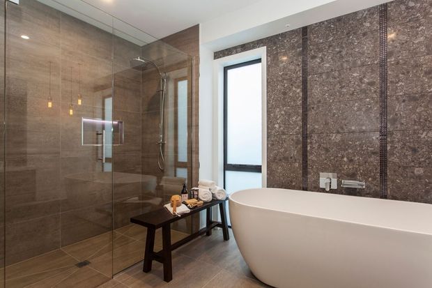 Luxurious contemporary bathroom featuring a large frameless glass walk-in shower, freestanding oval bathtub, narrow vertical window, dark stone-look wall cladding with contrasting textured strips, and warm brown tiled flooring. A dark timber bench with rolled towels and toiletries sits between the shower and bath.