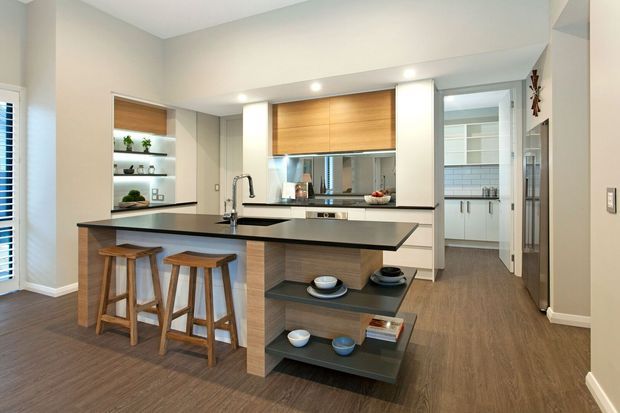 Contemporary open-plan kitchen with a large central island, black stone benchtop, integrated sink and tap, timber cabinetry, open shelving, and two timber bar stools. The kitchen opens toward a tiled utility or laundry area, with dark timber-look flooring and a neutral palette of white, beige, grey, and warm wood tones.