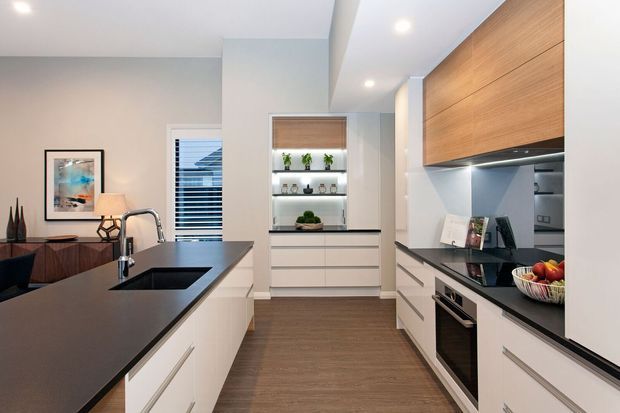 A bright, contemporary galley-style kitchen with a large dark stone island, white handleless cabinetry, integrated stainless-steel appliances, warm timber overhead cabinetry, and a black-framed display niche. Recessed ceiling lighting, pale walls, timber flooring, and a partially visible dining area create a clean, refined, functional interior.