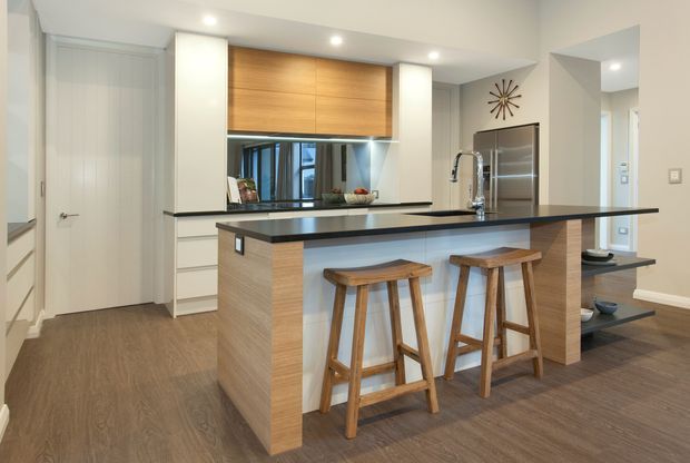 Contemporary kitchen with a large island and breakfast bar, featuring two timber stools, dark benchtops, light wood cabinetry, integrated appliances, recessed ceiling lights, and a reflective splashback. The kitchen opens toward a hallway and adjacent living area.