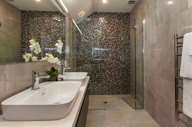 Contemporary bathroom with a long double vanity featuring white vessel basins and chrome tapware, a large wall mirror, glass-enclosed walk-in shower, and a striking dark patterned mosaic tile feature wall. Large light beige-grey tiles line the remaining walls and floor, with a heated towel rail and recessed ceiling lighting.