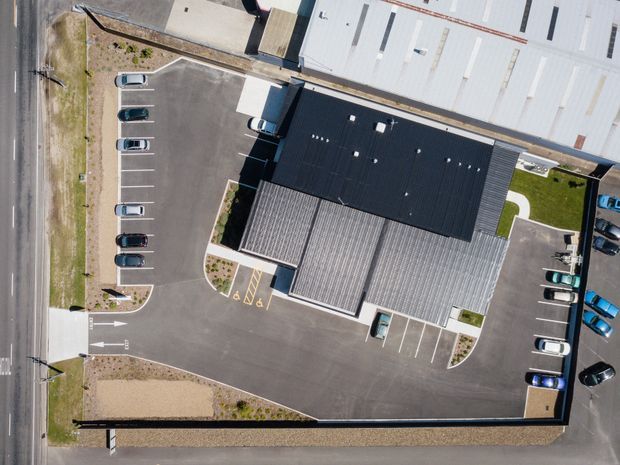 Top-down aerial view of a large commercial or industrial building with contrasting black and light grey roof sections, surrounded by paved parking areas, marked accessible spaces, driveways, planted edges, and adjacent roads. Several parked vehicles are visible around the site.
