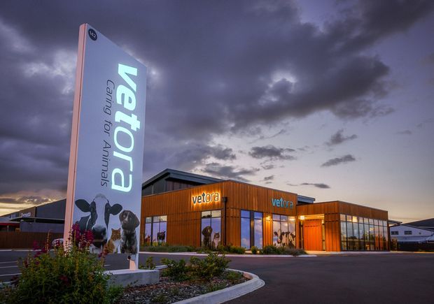 Exterior dusk photograph of a contemporary low-rise commercial animal-care building branded “Vetora Caring for Animals.” The building has warm timber-toned vertical cladding, extensive ground-level glazing, a projecting flat roof, illuminated signage, landscaped planting, and a paved driveway beneath a dramatic cloudy sky.