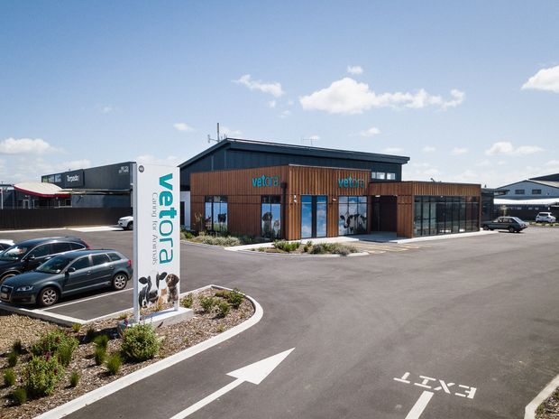 Exterior view of a contemporary low-rise commercial building in a paved parking and access area. The building has dark metal cladding, warm timber-look vertical wall cladding, extensive floor-to-ceiling glazing, and a projecting flat roofline. A prominent veterinary signage pylon and directional road markings identify the premises as a veterinary clinic or similar commercial facility. Landscaping is limited to low planted beds around the parking area.