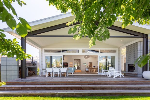 A bright contemporary outdoor entertaining area viewed from the garden, featuring a large covered terrace with a high single-slope roof, exposed dark structural beams, extensive white-framed glazing, broad timber steps and decking, an outdoor kitchen or barbecue at left, and white outdoor seating. The open doors reveal a dining and living area inside, while mature greenery frames the scene.
